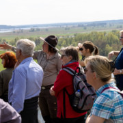 Danke-Exkursion für Naturwacht-Freiwillige Eine Gruppe von ca. 10 Erwachsenen schaut auf eine vor ihnen liegende Wiesenlandschaft mit Wasserflächen. Eine Rangerin weist mit der Hand in die Ferne.